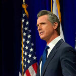 Man speaking at podium with American flags behind him.