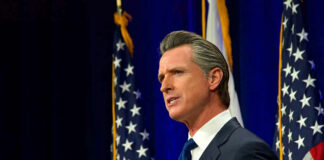 Man speaking at podium with American flags behind him.
