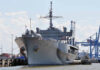 How One Warship Became A Budget Disaster Large naval ship docked at an industrial port.