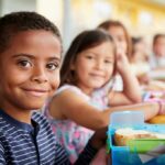 Farm-to-Tray Push Rattles Districts Smiling boy at a lunch table with classmates