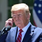 “America First” Just Hit an Identity Crisis man in suit at podium adjusting earpiece