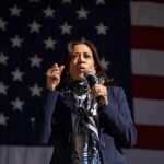Harris Praises Big-Government Icons — Not Biden woman speaking into a microphone in front of a large American flag