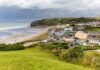 Emergency Powers Blitz As Surf Builds Coastal village by a wide sandy beach under cloudy skies