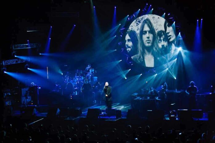 A band performing live on stage with dramatic lighting and a large screen displaying images of band members