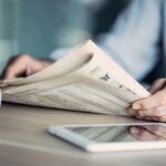 Hands holding a newspaper beside a tablet and coffee mug