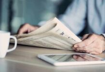 Fact-Check Torches Viral Migrant Claim Hands holding a newspaper beside a tablet and coffee mug