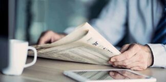 Hands holding a newspaper beside a tablet and coffee mug
