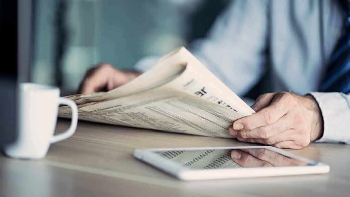 shutterstock_1438272785 Hands holding a newspaper beside a tablet and coffee mug