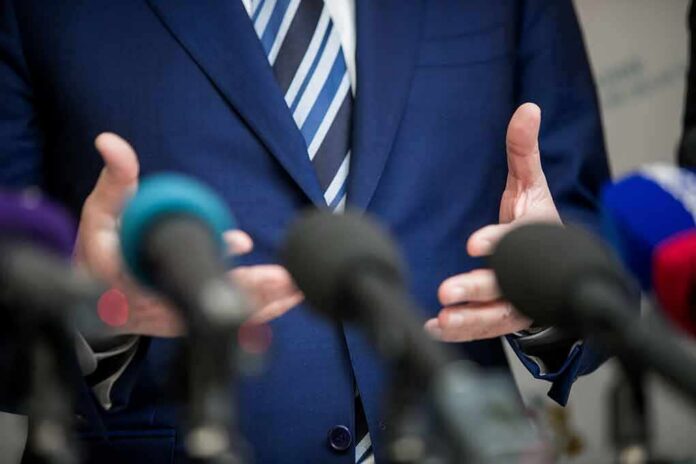 shutterstock_1670760397 Speaker in blue suit gesturing behind microphones