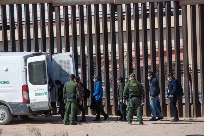 shutterstock_2079408475-1 Border agents escort a group near a tall fence and a patrol van