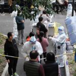 Fast-Spread Ebola Shatters Records Christian Jones and others wait at an outdoor COVID-19 testing site with staff in protective suits