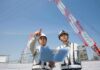 Two construction workers reviewing plans at a site with cranes