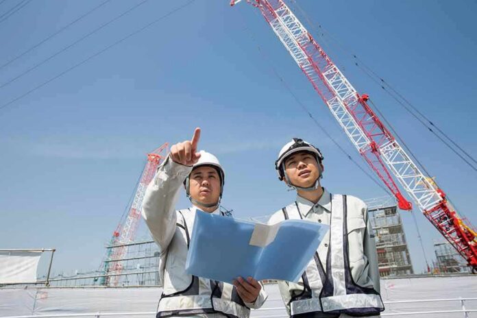 shutterstock_2154192201 Two construction workers reviewing plans at a site with cranes