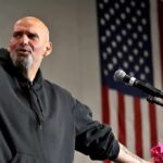 Bald man speaking at a microphone with a U.S. flag in the background