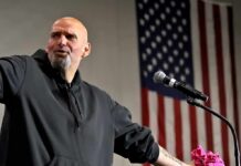 Bald man speaking at a microphone with a U.S. flag in the background