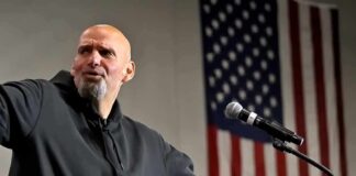 Bald man speaking at a microphone with a U.S. flag in the background