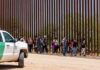 Caravan Tracked North – Crackdown Looms Group of people walking along a tall border fence beside a patrol vehicle