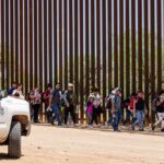 Caravan Tracked North – Crackdown Looms Group of people walking along a tall border fence beside a patrol vehicle