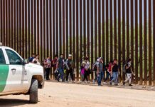Group of people walking along a tall border fence beside a patrol vehicle