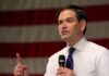 Rubio Consolidates Power – Two Hats, One Boss man speaking with microphone in front of an American flag