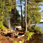 Tent and campfire by a forested lakeshore at sunset