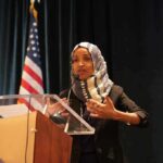 Ilhan Omar’s Home is Raided by Feds! Ilhan Omar speaking at a podium with a microphone