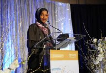 Denaturalization Threat Looms Over Omar Ilhan Omar speaking at a podium on a decorated stage