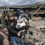 Rescuers carry a baby in a stroller through a destroyed bridge