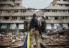 Deadly Strikes Rattle Capital Soldier with rifle stands before a destroyed building holding a blue and yellow flag