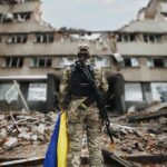 Soldier with rifle stands before a destroyed building holding a blue and yellow flag