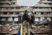 Soldier with rifle stands before a destroyed building holding a blue and yellow flag