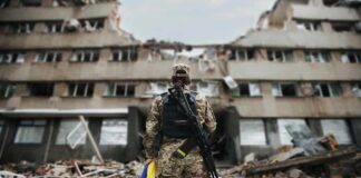 Soldier with rifle stands before a destroyed building holding a blue and yellow flag