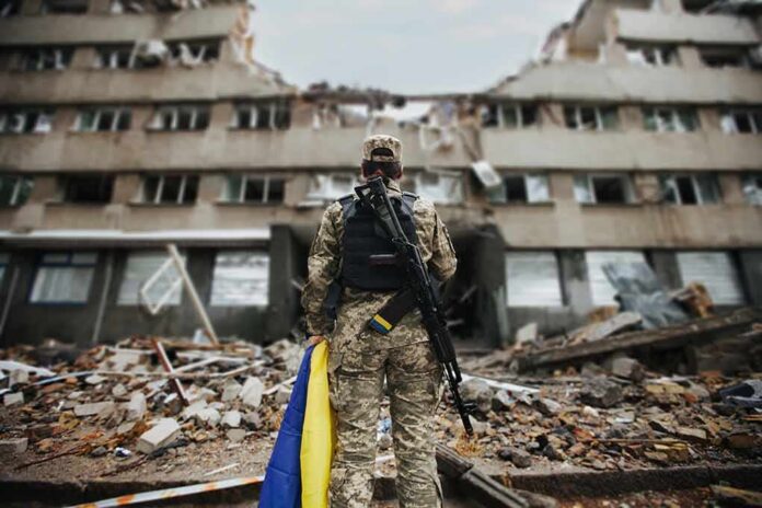 Soldier with rifle stands before a destroyed building holding a blue and yellow flag