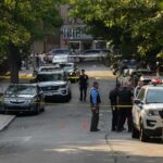 Homecoming Horror – Dozens Down, Killers Loose Police officers secure a residential street with patrol cars and yellow tape