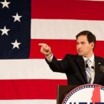 Speaker in suit pointing during speech before large American flag