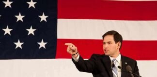Speaker in suit pointing during speech before large American flag