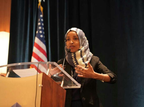 Woman speaking at podium with American flag background.