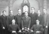 Group portrait of men associated with the Skull and Bones secret society, featuring a skull and crossed bones on a table