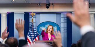 Press secretary at podium, answering reporters questions, raised hands.