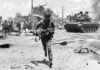 A soldier running through a war-torn area with a tank in the background