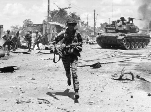 A soldier running through a war-torn area with a tank in the background
