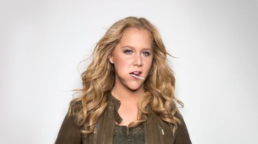A woman with blonde hair holding a lollipop in her mouth, posing confidently against a plain background