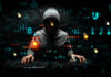 Hooded hacker using two keyboards surrounded by code.