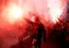 Protesters with flares walk through smoky urban street.