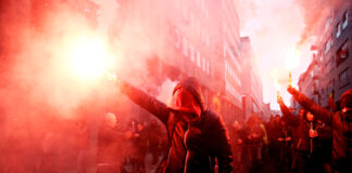 Protesters with flares walk through smoky urban street.