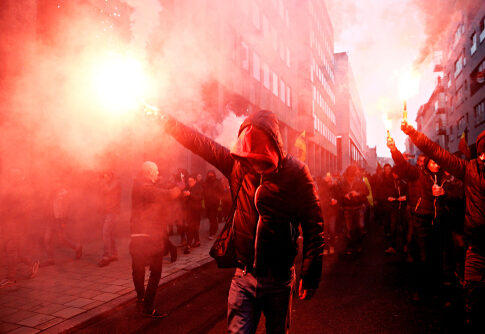 Protesters with flares walk through smoky urban street.