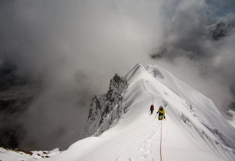 Climbers ascending a snowy mountain ridge under a cloudy sky