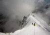 Climbers ascending a snowy mountain ridge under a cloudy sky