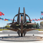 NATO emblem with flags of member countries in background.