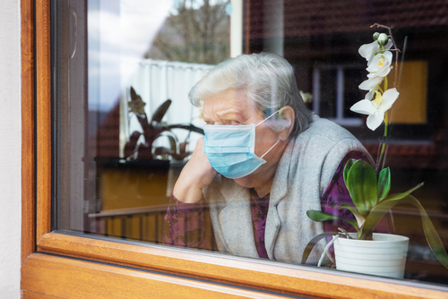 shutterstock_1688780245.jpg featured image Elderly woman wearing mask, looking out window beside orchid.